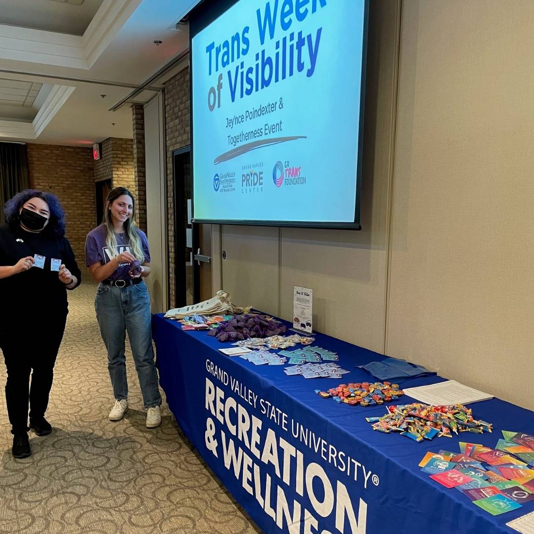 Image of two RecWell staff by a table full of fun wellness supplies in front of "Trans Week of Visibility" projected on a screen